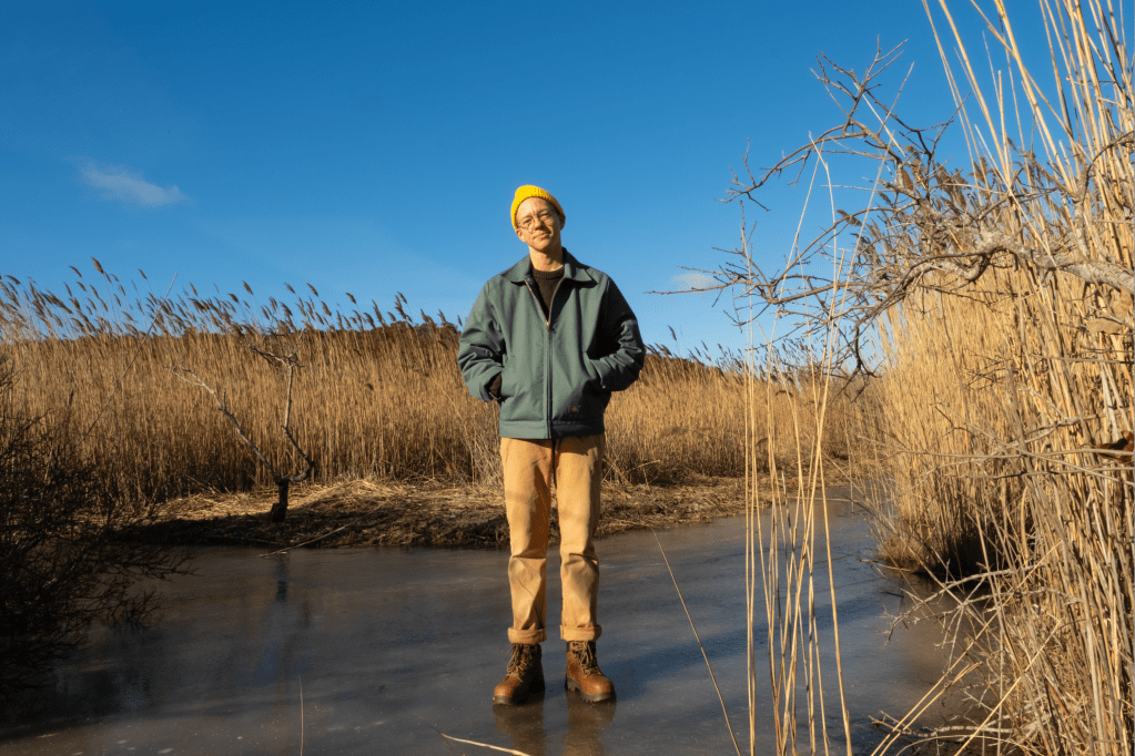 Acie wears a jacket and a yellow hat, standing on a frozen pond, surrounded by reeds.